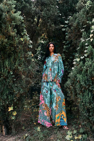 Cinched-waist shirt with Gond art–inspired placement prints, pearl embroidery, shell tassel details, and open-back tie-up design, paired with matching printed crepe trousers featuring a flared silhouette and side slit. Designed by Limerick.