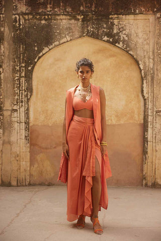 A model posing in a resort look. The dress is in coral colour made from linen crop top, Linen jacket with cut work floral embroidery, silk organza warp pre-stitched skirt. A perfect resort look book dress, holiday, vacation, coastal, beach wear.