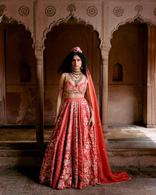 A red multi-tone lehenga, featuring a delightful combination of leheriya and ornamental signature floral prints that evoke the charm of the ancient craftsmanship of Rajasthan. It is paired with an intricate hand–embroidered strappy blouse, and a delicate hand–embroidered scallop dupatta.